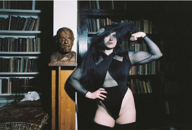 Woman poses indoors wearing black bodysuit and hat with veil, showcasing biceps, with books and sculpture in the background.