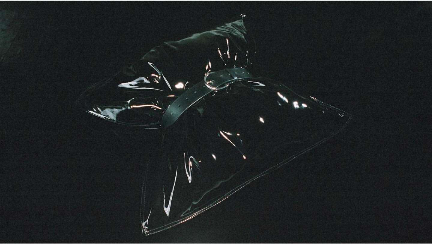 Clear, vinyl fashion accessory item with adjustable belt and metallic buckle, laying against a dark background with visible lighting.