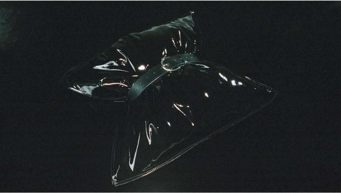 Clear, vinyl fashion accessory item with adjustable belt and metallic buckle, laying against a dark background with visible lighting.