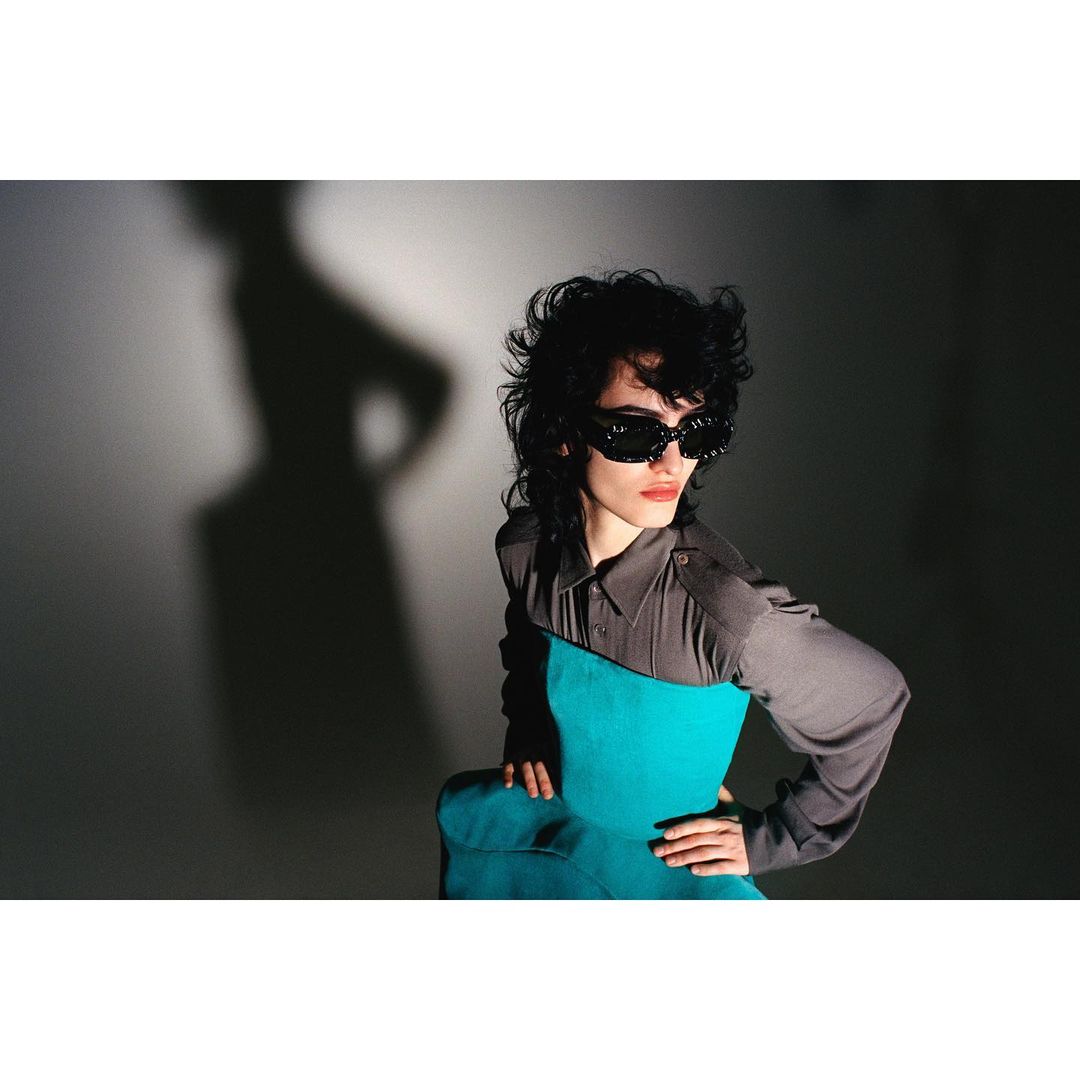 Fashionable woman in sunglasses posing indoors. She is wearing a turquoise top with a grey collared shirt and has dark curly hair.