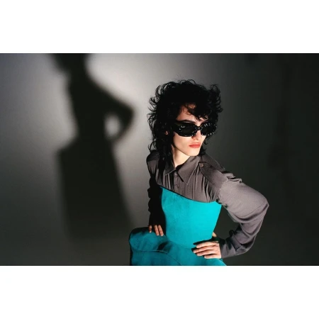 Fashionable woman in sunglasses posing indoors. She is wearing a turquoise top with a grey collared shirt and has dark curly hair.
