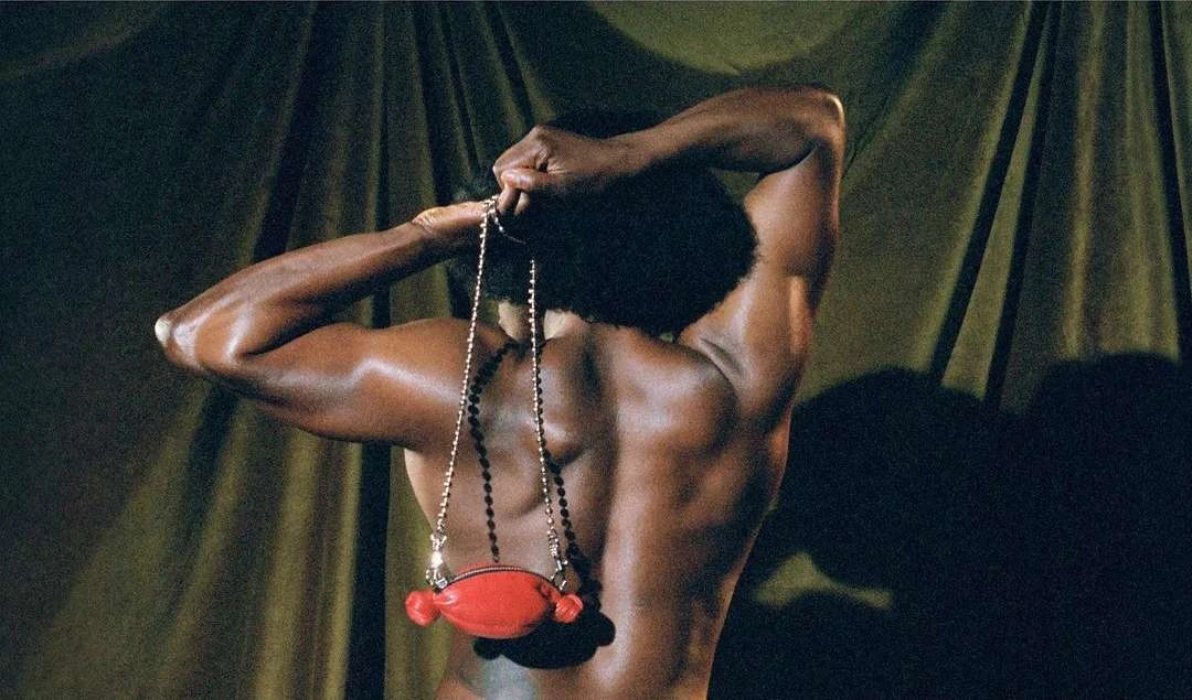 View of a Black adult male from the back, displaying strong musculature and holding a red purse with beaded strap against a green drapery backdrop.