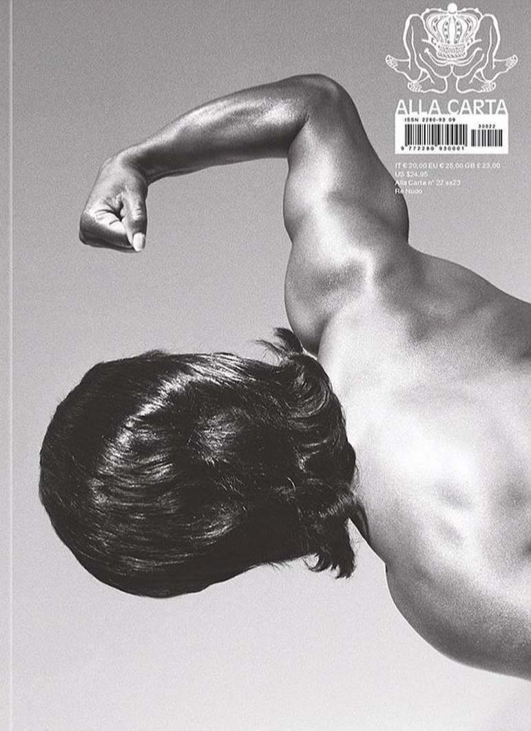 Monochrome photograph of a muscular adult with long dark hair posing shirtless in a grey-toned setting, showcasing body physique.