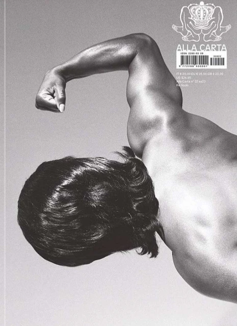 Monochrome photograph of a muscular adult with long dark hair posing shirtless in a grey-toned setting, showcasing body physique.