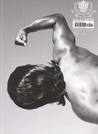 Monochrome photograph of a muscular adult with long dark hair posing shirtless in a grey-toned setting, showcasing body physique.