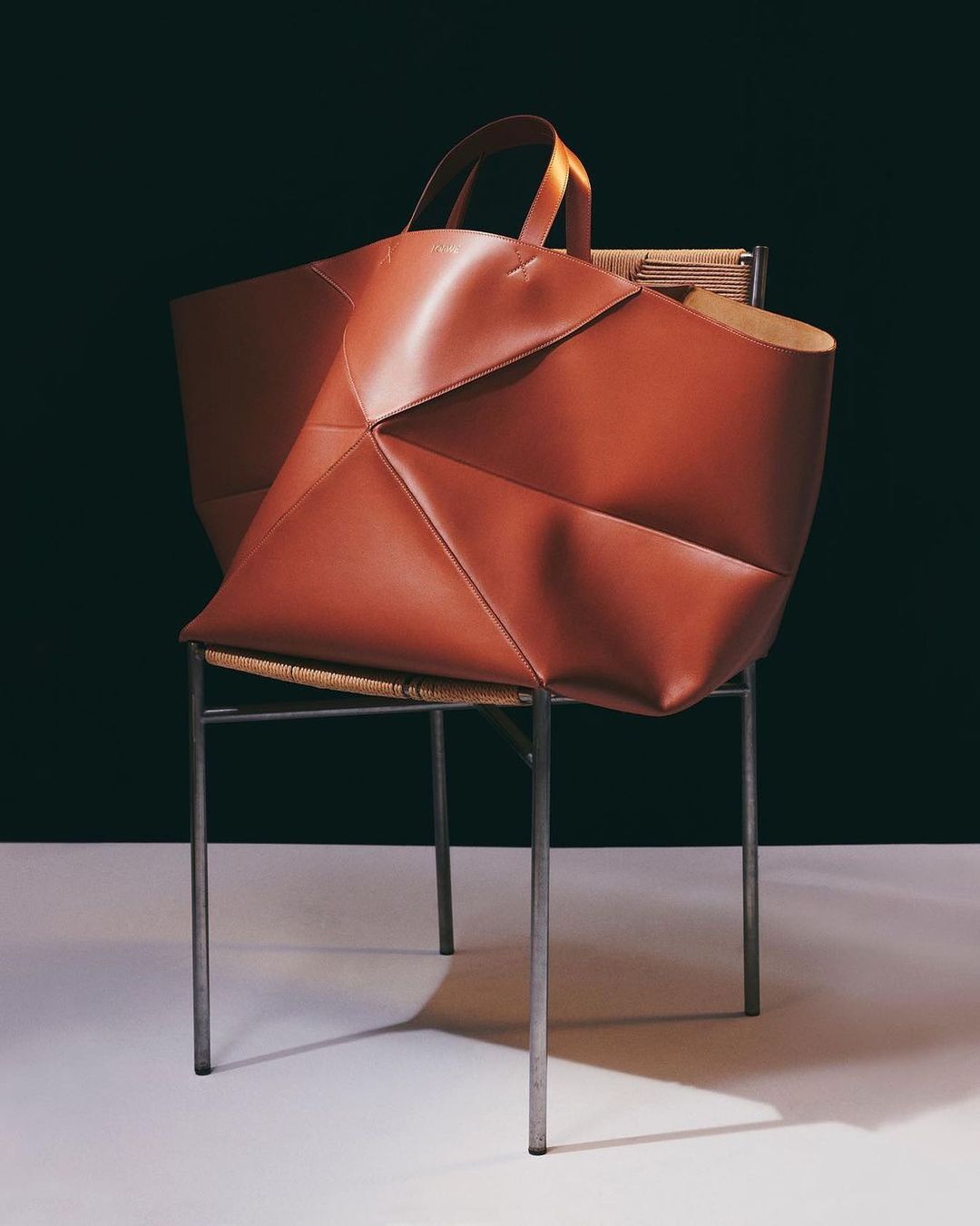 Contemporary leather tote bag in reddish-brown on woven-seat chair against a dark background in a studio fashion product shot.