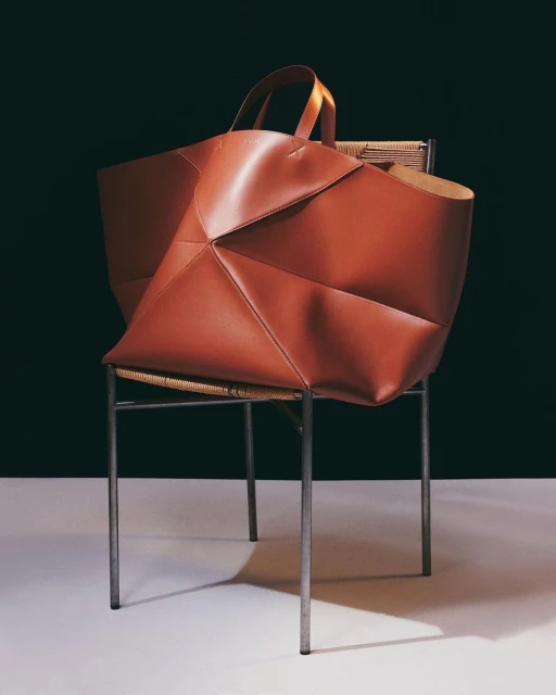 Contemporary leather tote bag in reddish-brown on woven-seat chair against a dark background in a studio fashion product shot.