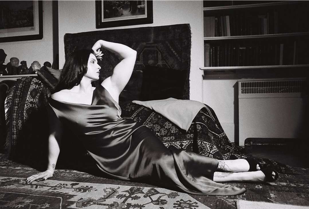 Woman in a long dark slip dress reclining on a patterned rug in a domestic living room by Camille Vivier
