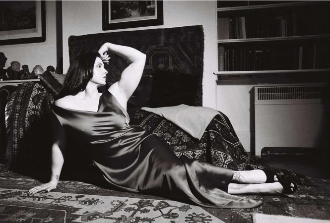 Woman in a long dark slip dress reclining on a patterned rug in a domestic living room by Camille Vivier