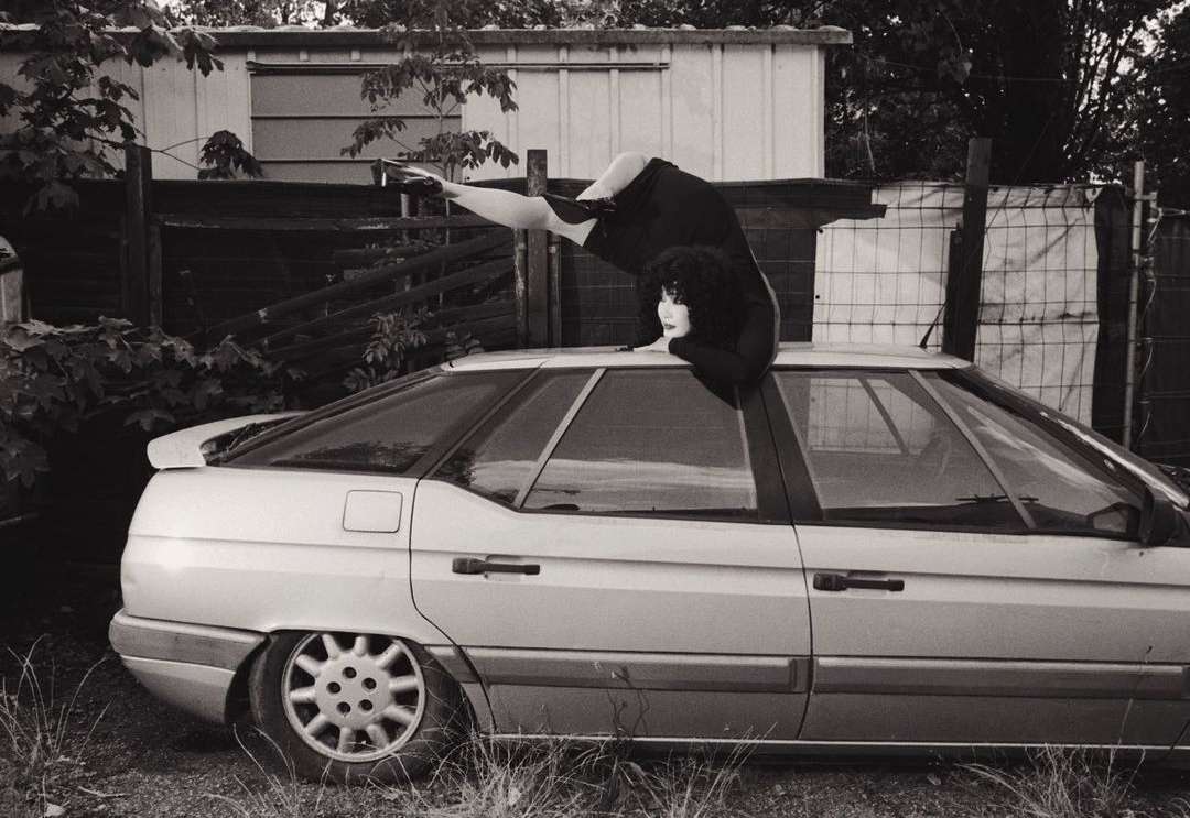 Monochrome image of a woman on a silver car, one leg raised, with a metal fence and a white building in the background, shot at eye-level.