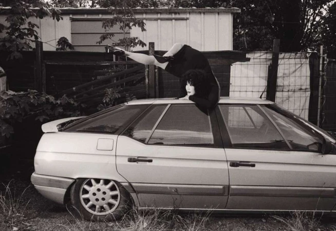 Monochrome image of a woman on a silver car, one leg raised, with a metal fence and a white building in the background, shot at eye-level.