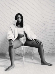 Black and white portrait of a woman seated on a small chair wearing a puffer jacket and cut out bodysuit.