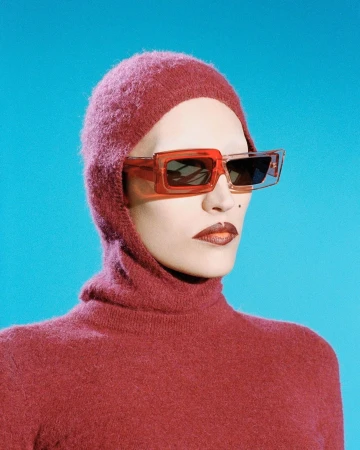 Model wearing a pink knitted balaclava and transparent red sunglasses against a bright blue background by Camille Vivier.