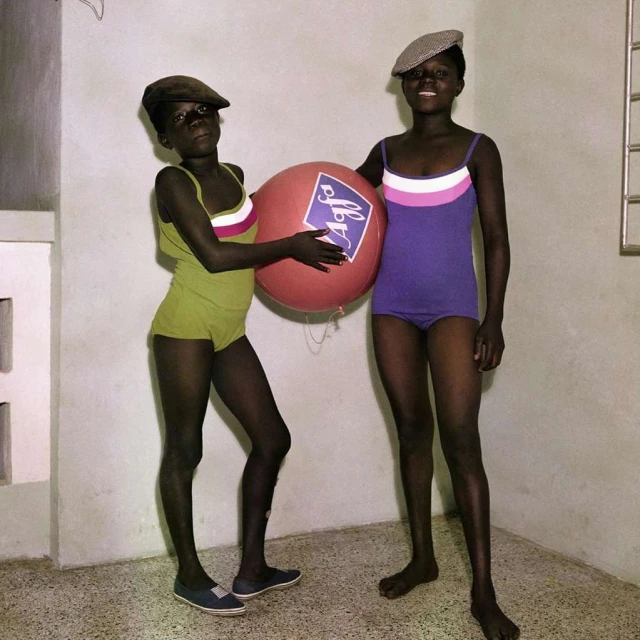 Two children in swimsuits stand beside an Agfa-branded red balloon in an indoor setting, wearing matching hats.