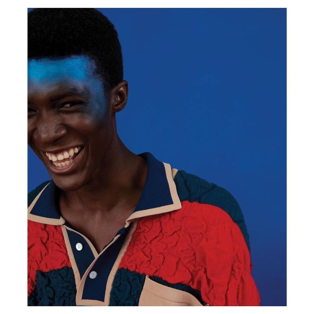 Portrait of a Black man smiling with blue face paint, wearing a colorful polo shirt, against a solid blue background.