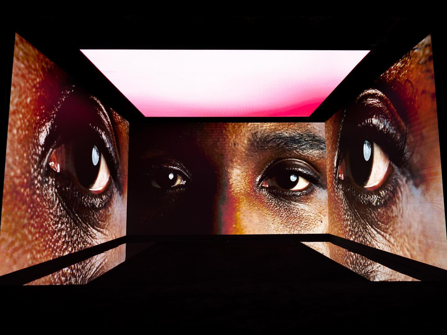 A close-up of a person's face is projected onto three walls, focusing on the eyes, set against a black backdrop with pink and white lighting.