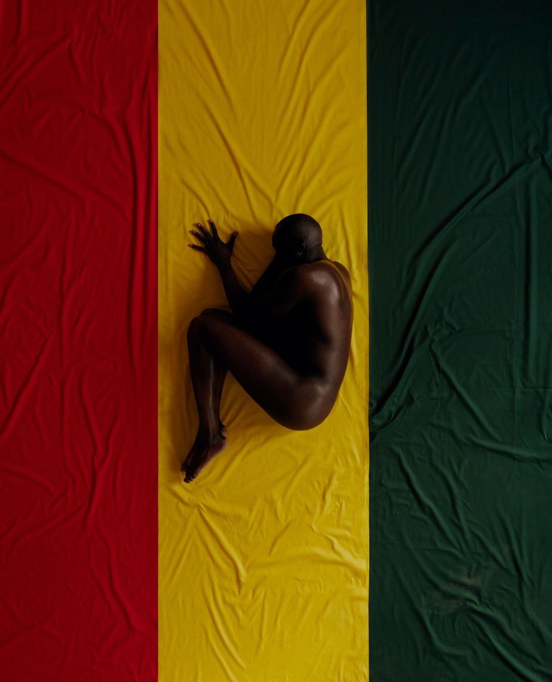 Nude adult male lies curled on a textile colored red, yellow, and green in a studio setting. Striking composition.