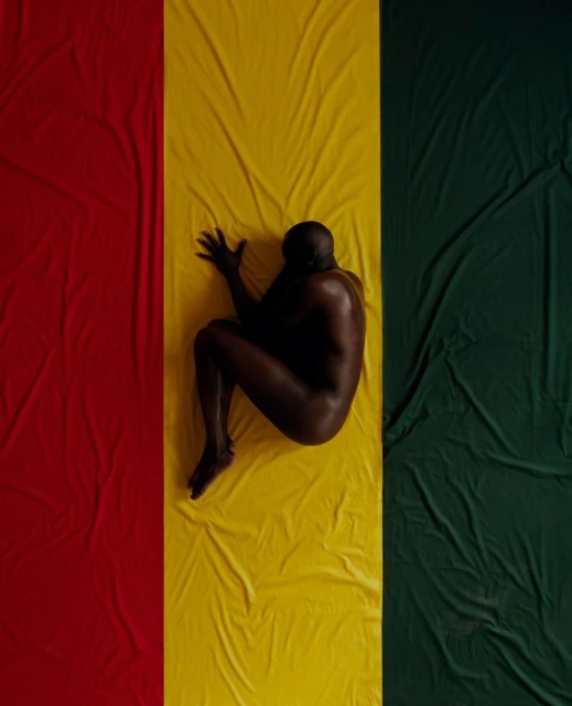 Nude adult male lies curled on a textile colored red, yellow, and green in a studio setting. Striking composition.