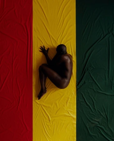 Nude adult male lies curled on a textile colored red, yellow, and green in a studio setting. Striking composition.