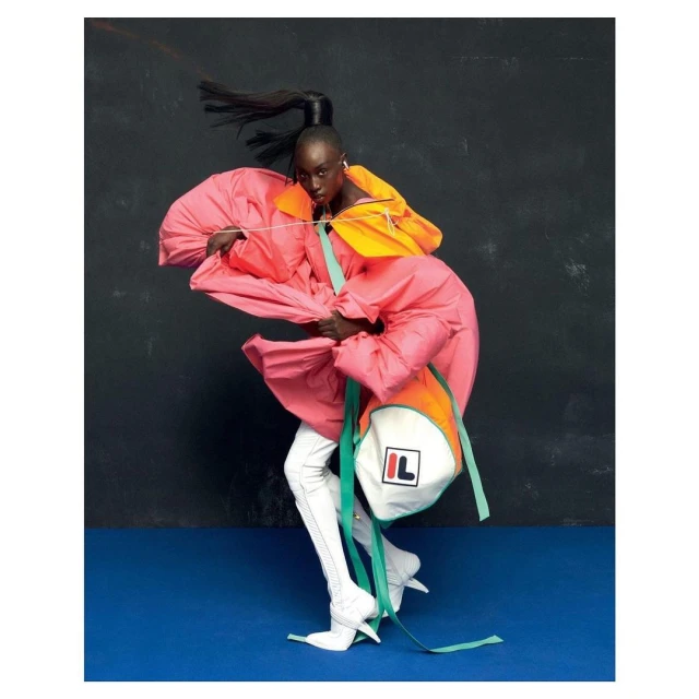 Fashion editorial shot of a Black woman in oversized pink and orange jacket, thigh high boots, and logo backpack against dark blue backdrop.