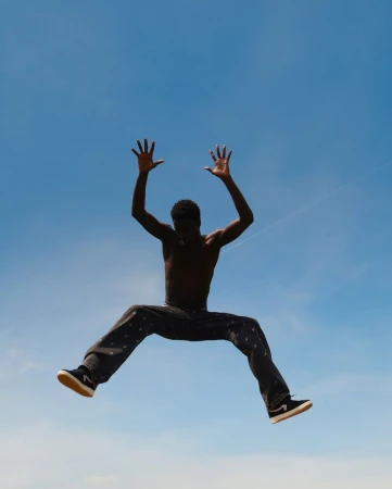 A shirtless black man captured mid-air jumping against a clear blue sky background.