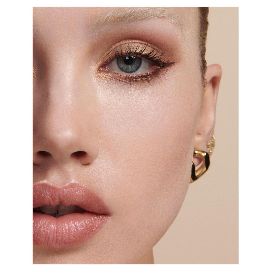 Close-up beauty shot of an adult female with makeup and gold earrings against a beige background in a studio setting.