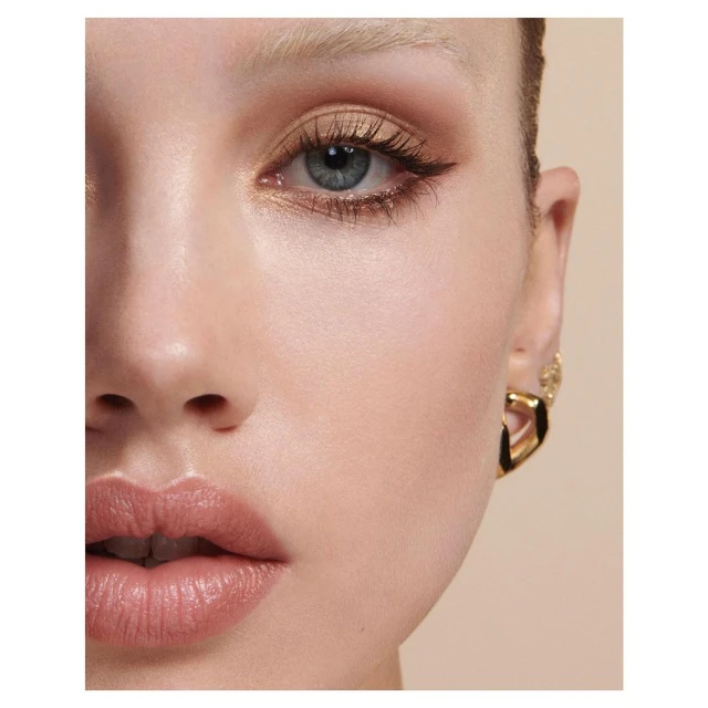 Close-up beauty shot of an adult female with makeup and gold earrings against a beige background in a studio setting.