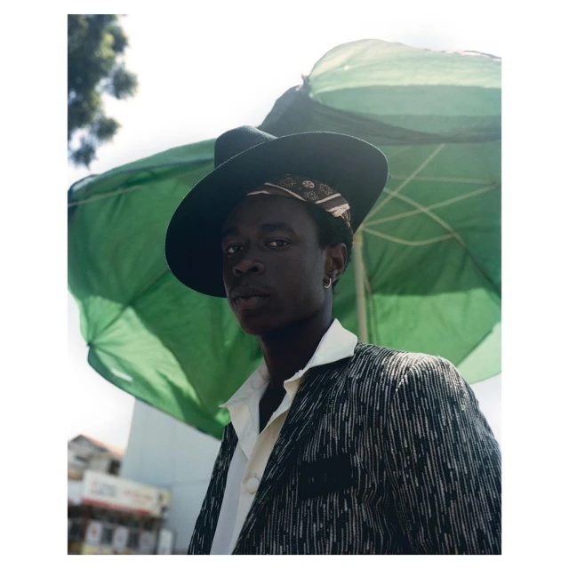 Stylish African man poses under green sun umbrella. He wears a patterned blazer, white shirt, hat, and earring in an outdoor setting.