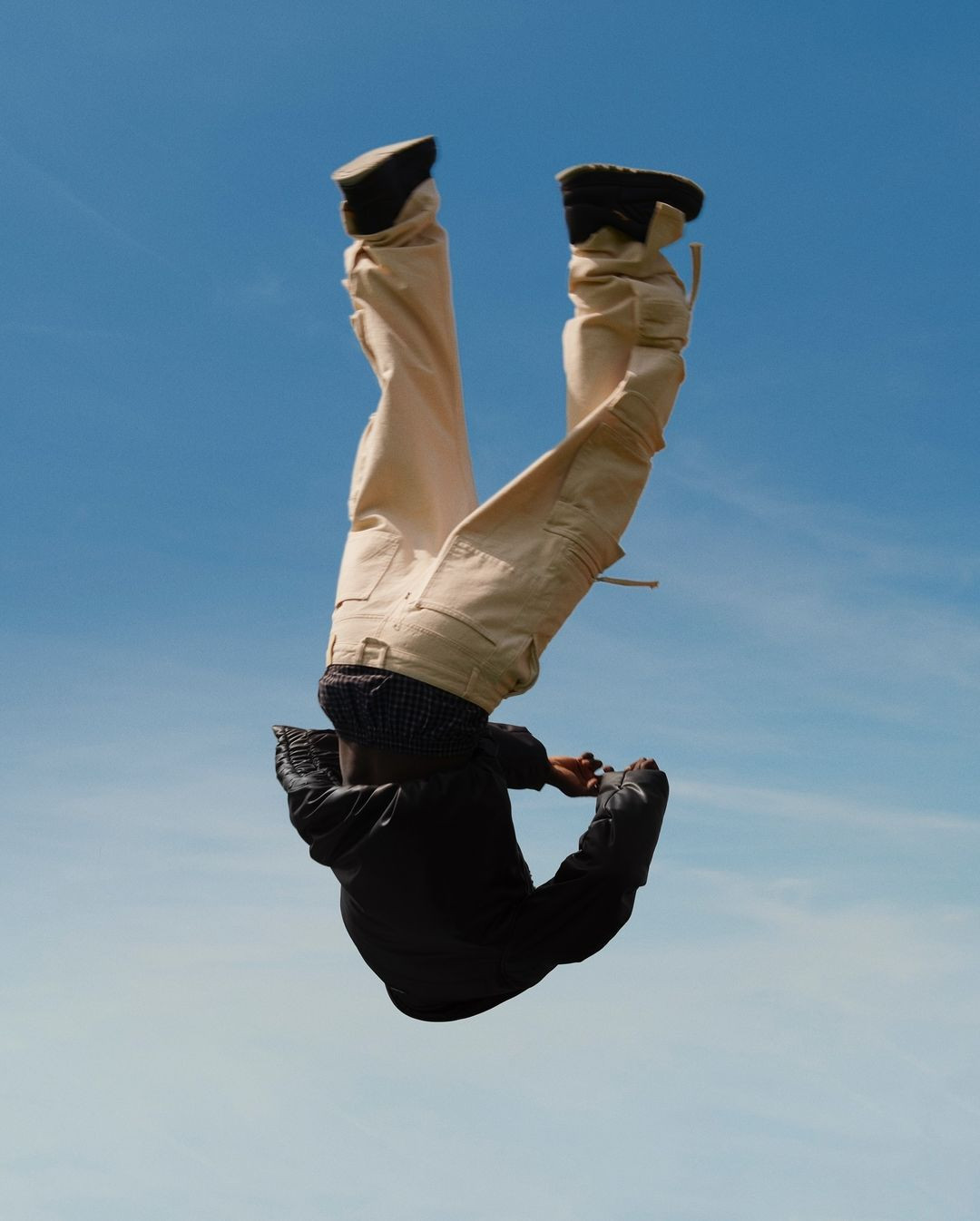 Person in mid-air wearing a black jacket, khaki pants, and black shoes against a blue sky with light clouds, inverted perspective.