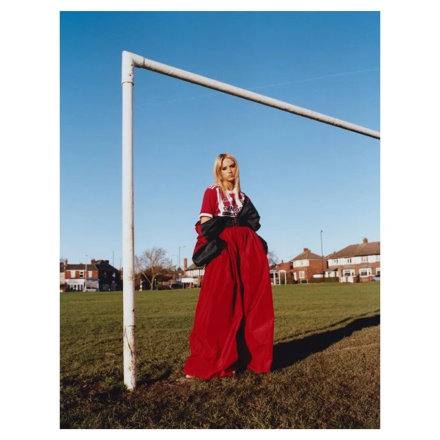 A blonde woman in a red dress and soccer jersey stands in a grassy field next to a soccer goal, with houses visible in the background.