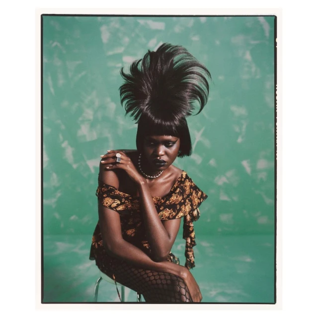 A dark-skinned woman with an avant-garde hairstyle sits on a stool, wearing a floral top and fishnet stockings, against a green backdrop.