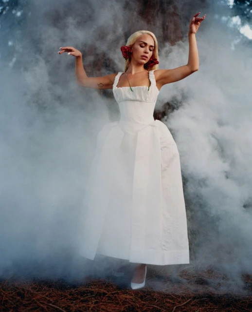 Woman in a white gown posing in a misty outdoor setting, with red floral hair accessories and white pointed heels.