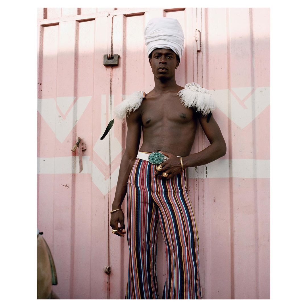 Portrait of an adult male wearing a white turban and vertically striped pants, standing in front of a pink wall with white accents.