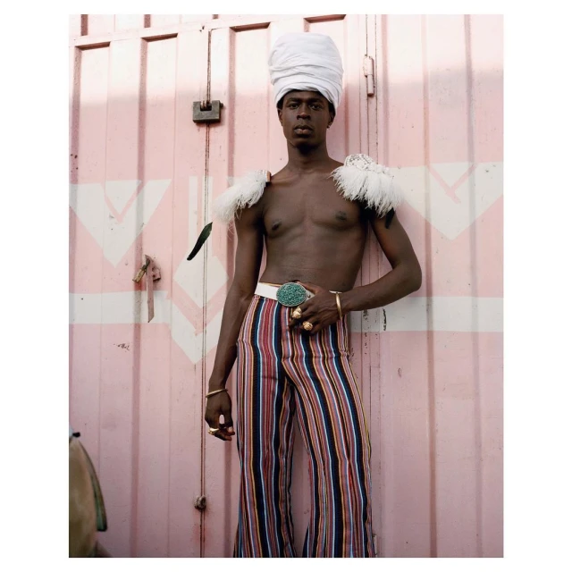 Portrait of an adult male wearing a white turban and vertically striped pants, standing in front of a pink wall with white accents.