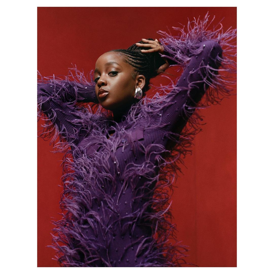 Portrait of an African American woman with braids, wearing a purple feathered dress, against a red background in a studio setting.