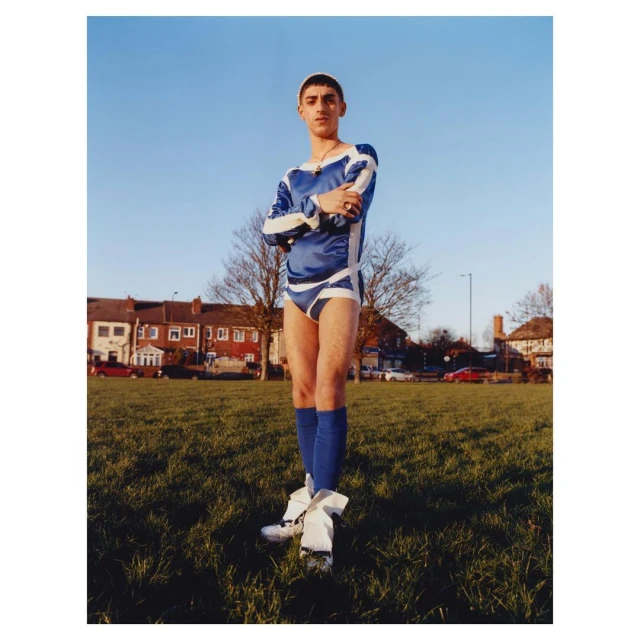 A young adult male stands on a grassy field wearing a blue and white soccer uniform with his arms crossed, posing outdoors on a sunny day.