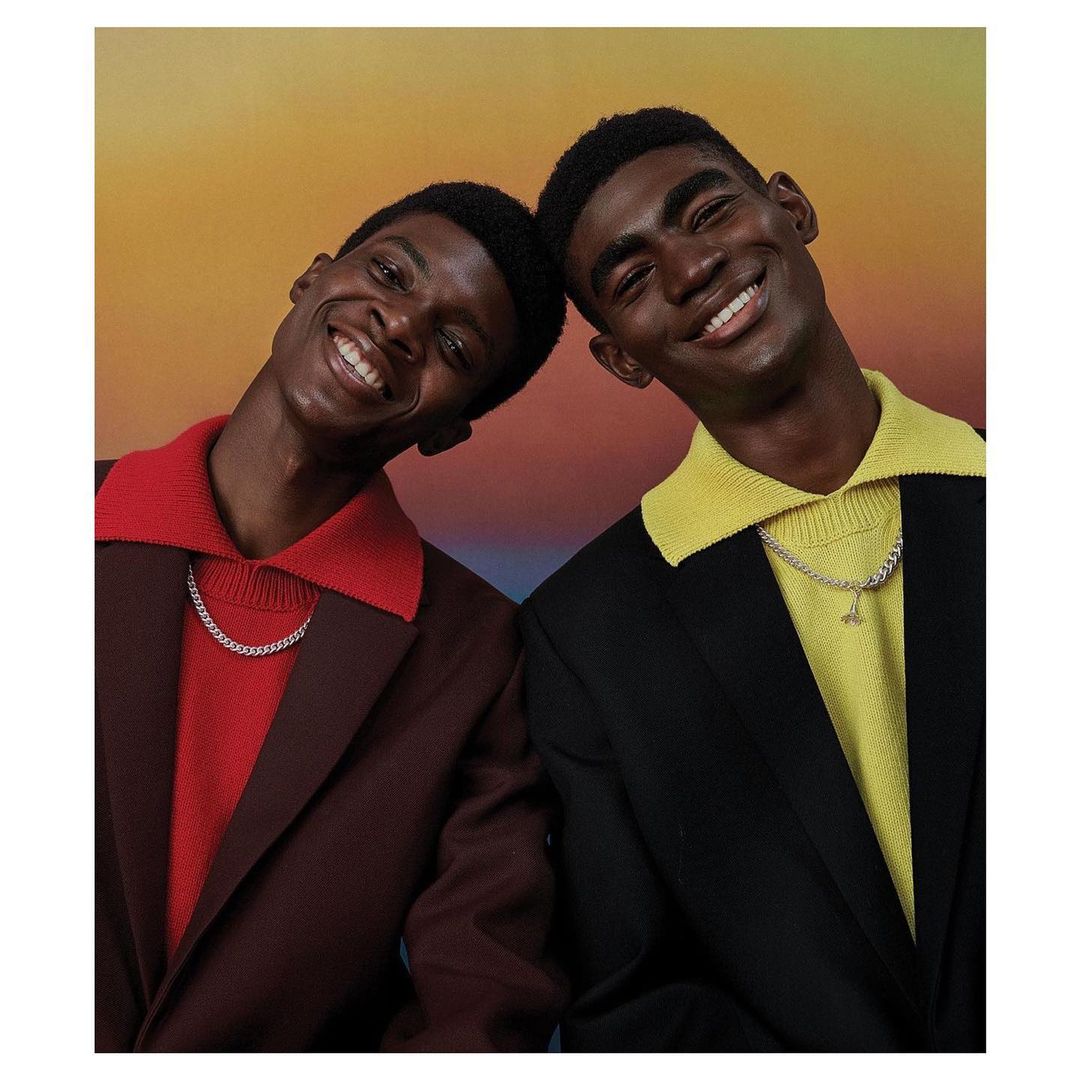 Two Black men in collared sweaters and suit jackets smile against a gradient background, wearing silver necklaces, portrait orientation.