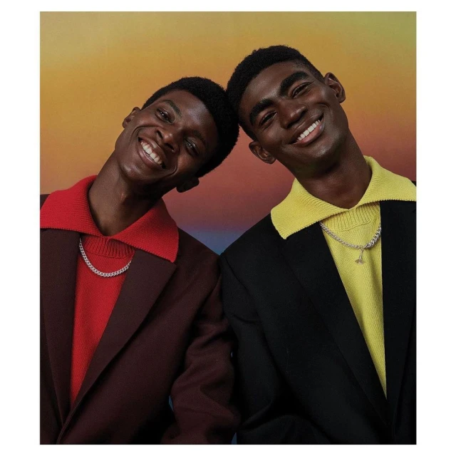Two Black men in collared sweaters and suit jackets smile against a gradient background, wearing silver necklaces, portrait orientation.