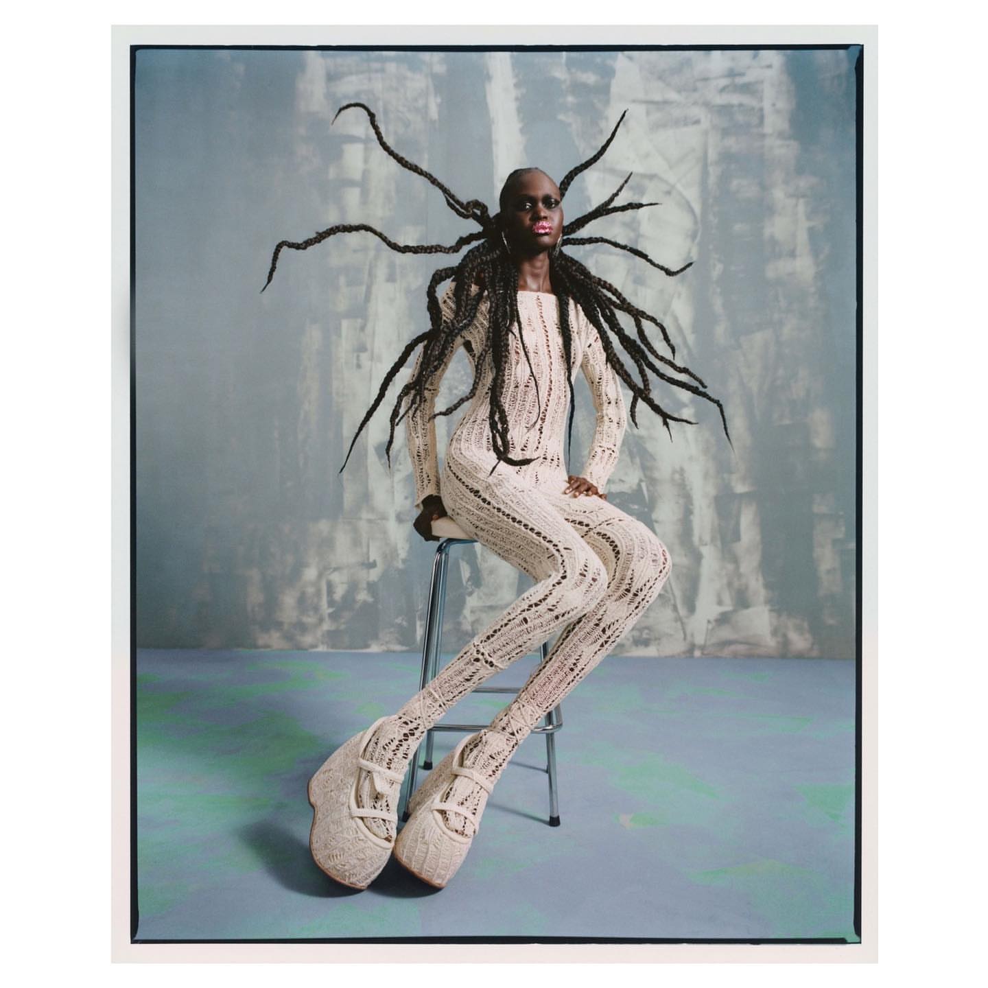 A person with dark skin and elaborate hair, seated on a metal stool, is wearing a cream-colored, patterned knit outfit and sculptural shoes against an abstract backdrop.