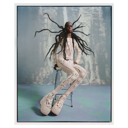A person with dark skin and elaborate hair, seated on a metal stool, is wearing a cream-colored, patterned knit outfit and sculptural shoes against an abstract backdrop.