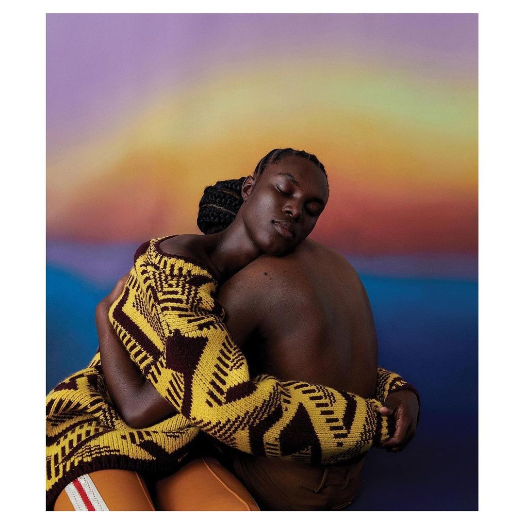 Studio portrait of two people embracing, one wearing a geometric-patterned sweater and the other bare-chested, against a gradient background.