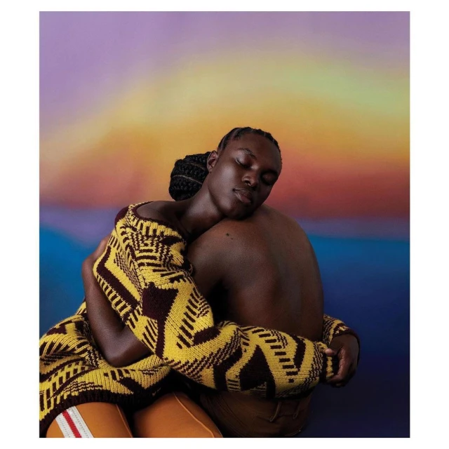 Studio portrait of two people embracing, one wearing a geometric-patterned sweater and the other bare-chested, against a gradient background.