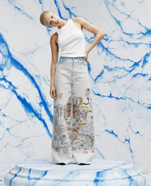 Adult female fashion model poses in white top and pants with graphic art print, against a faux marble background in a studio shot.