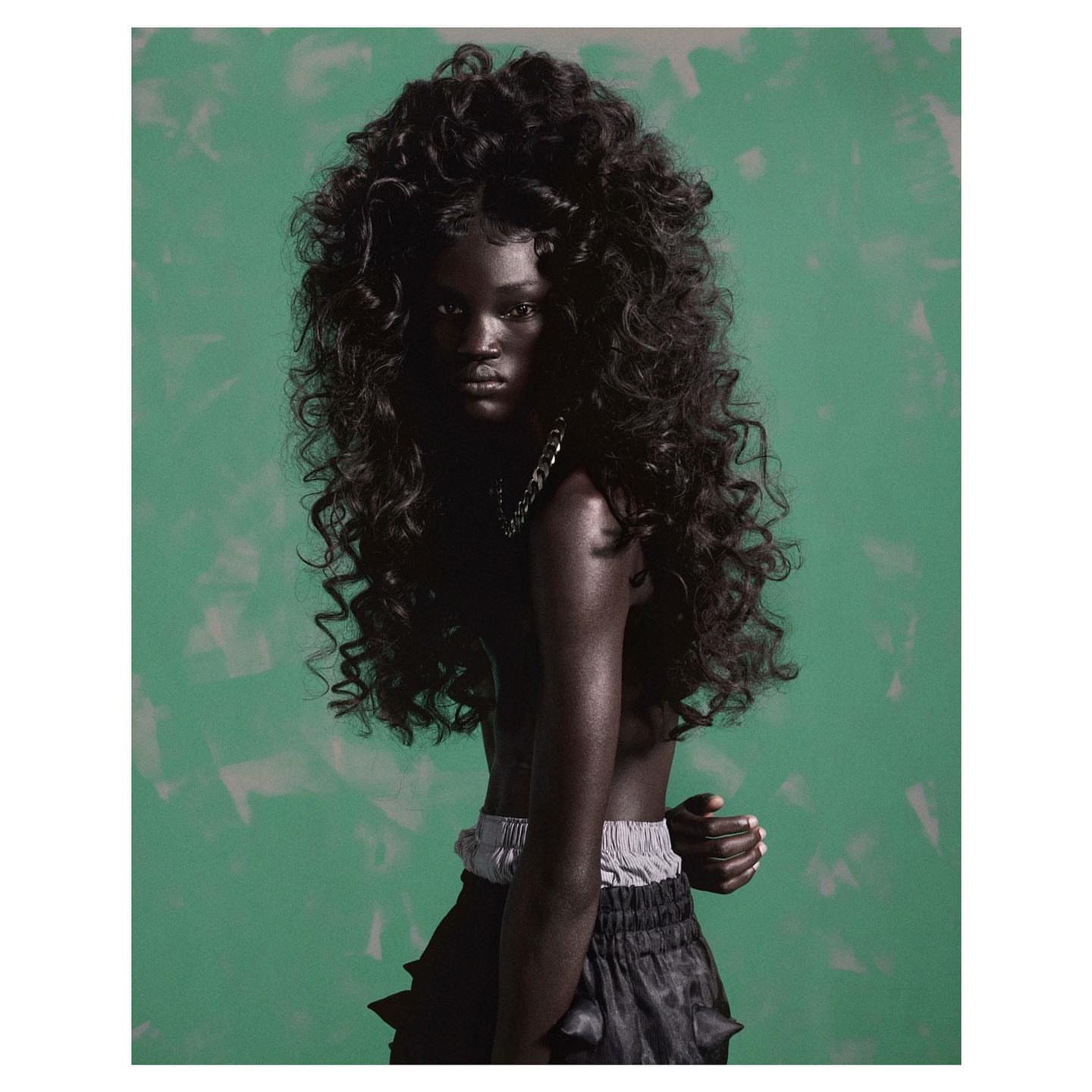 A striking close-up shot captures a dark-skinned woman with voluminous curly black hair, silver necklace, and a green, textured backdrop.