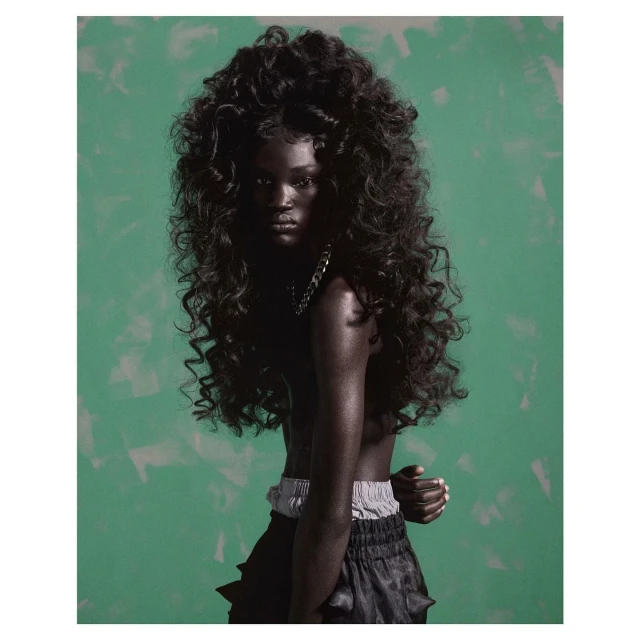 A striking close-up shot captures a dark-skinned woman with voluminous curly black hair, silver necklace, and a green, textured backdrop.