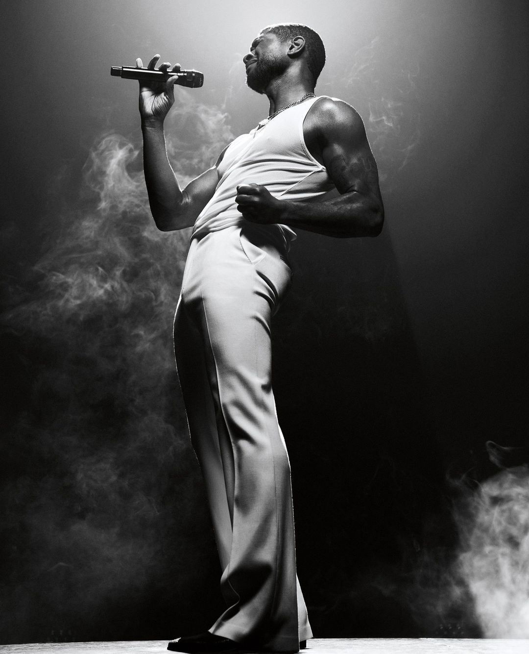 Monochrome portrait of a male singer in a tank top and wide-leg trousers, holding a microphone, on stage in a cloud of smoke.