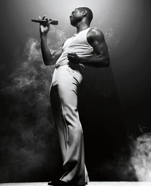 Monochrome portrait of a male singer in a tank top and wide-leg trousers, holding a microphone, on stage in a cloud of smoke.