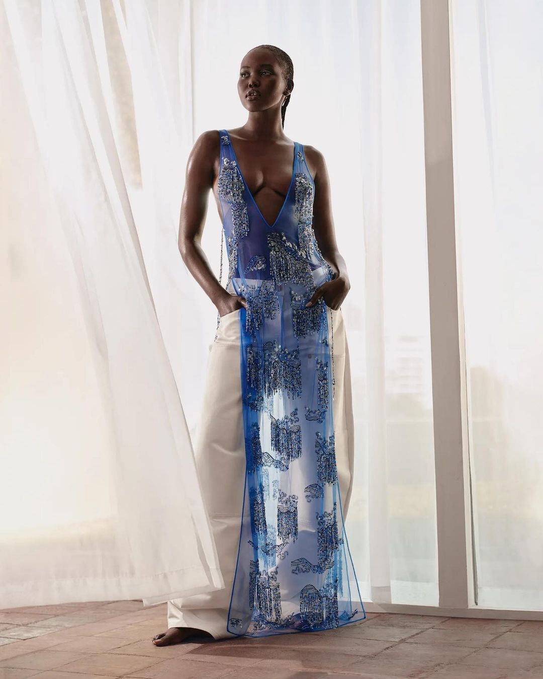 A fashionable dark-skinned woman posing in a sleeveless top and white pants in front of sheer white curtains and neutral background.