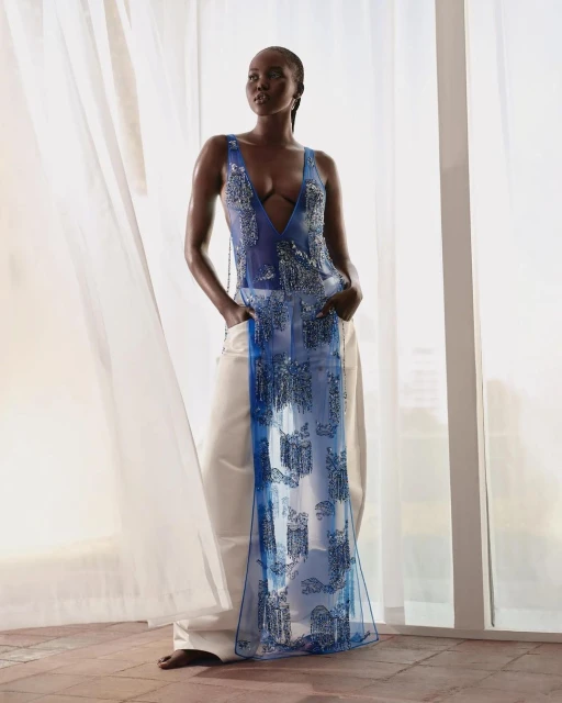 A fashionable dark-skinned woman posing in a sleeveless top and white pants in front of sheer white curtains and neutral background.