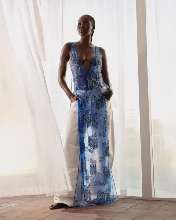 A fashionable dark-skinned woman posing in a sleeveless top and white pants in front of sheer white curtains and neutral background.